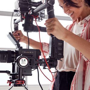 Female Videographer With Video Camera On Steady Cam Rig Filming Movie In White Studio